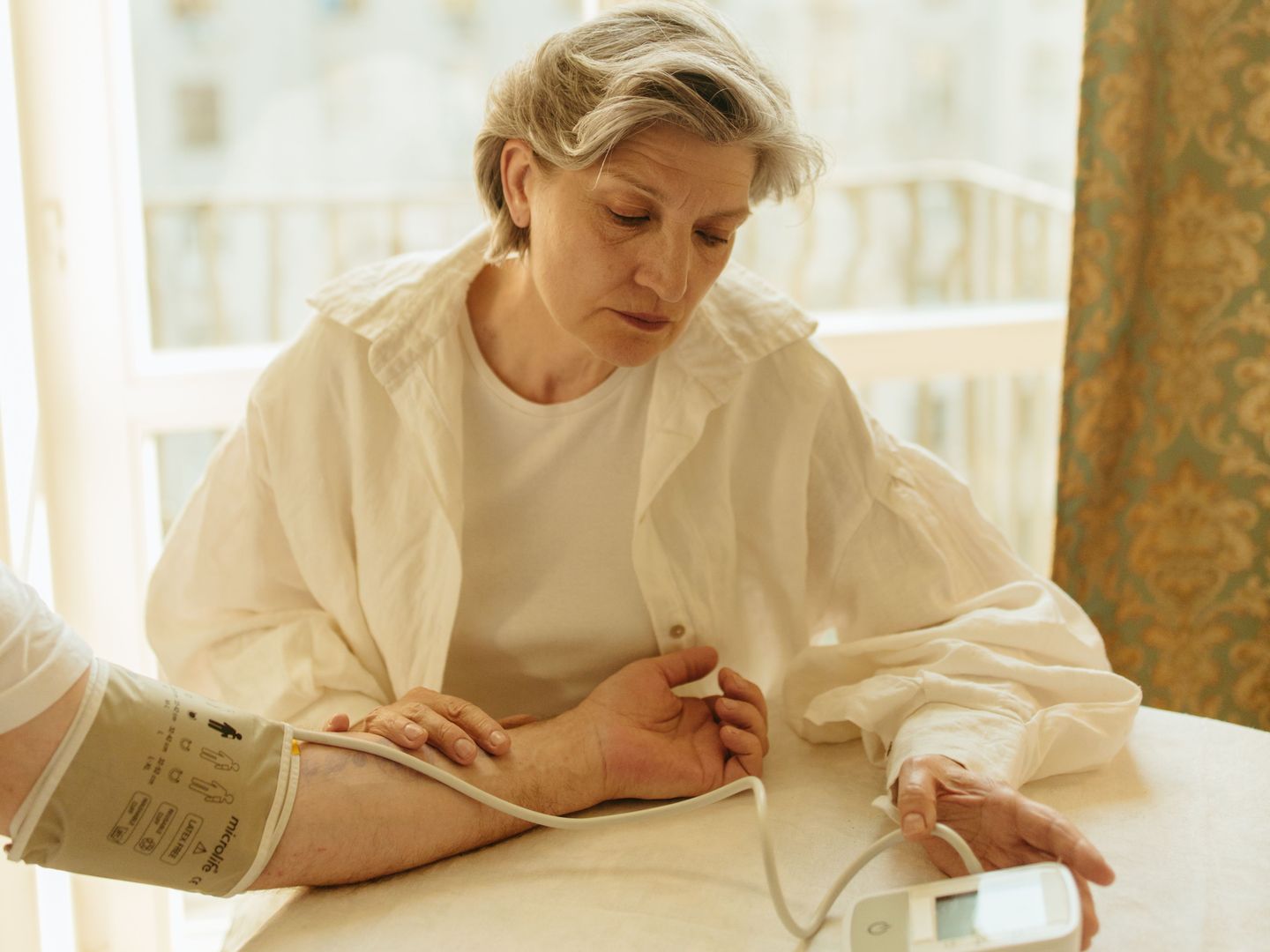 An older woman at a sunlit table at home, taking a blood pressure reading with a home monitor.